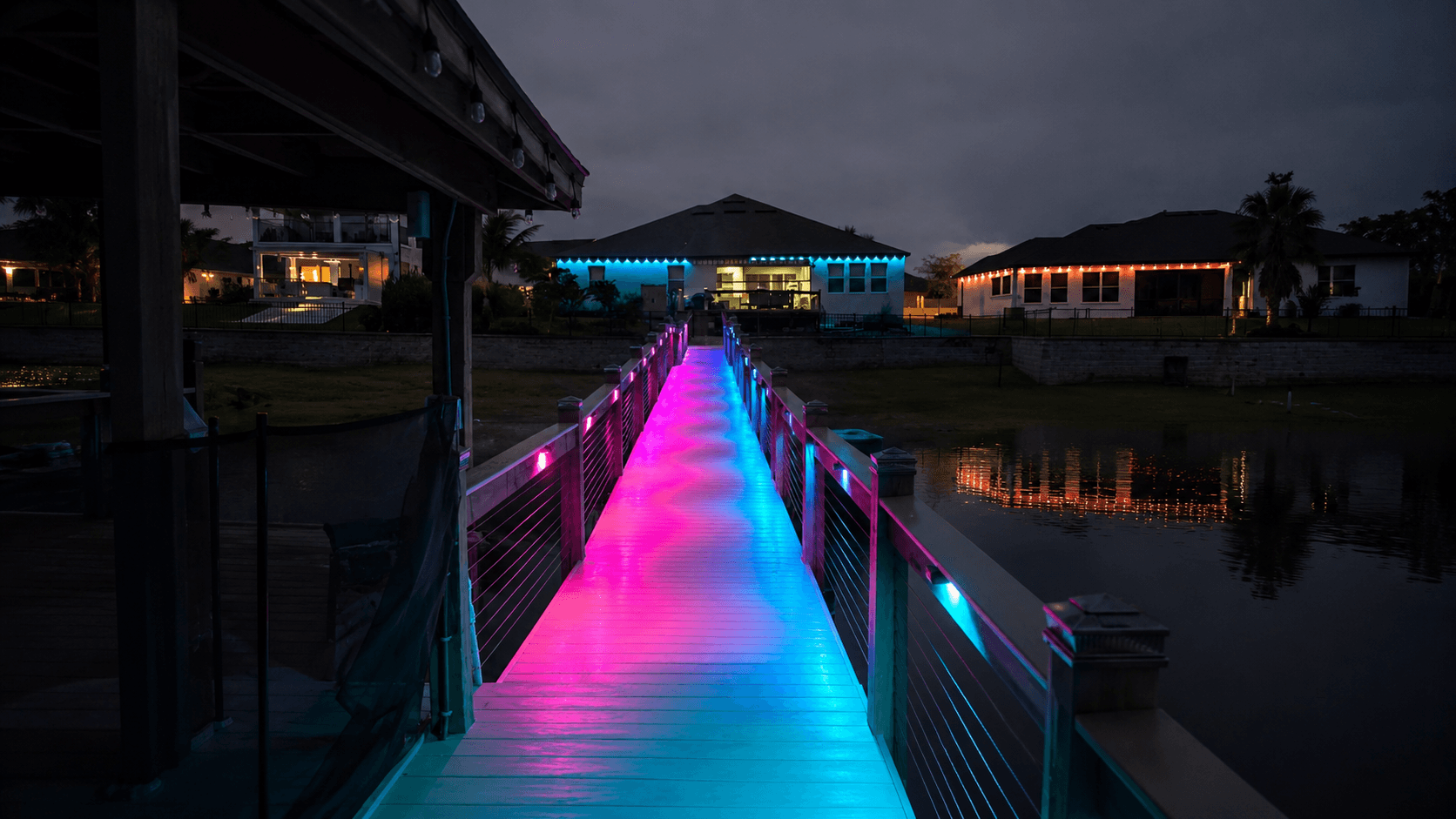 Illuminated dock at night with vivid pink and teal Govee RGBWIC lighting running along the walkway over a Florida waterway, with homes in the background also lit with smart color roofline lights